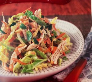 Big mound of Vietnamese chicken salad piled on a white plate with black trim on a red background, showing mint, carrots, and cabbage plus shredded hcicken breast