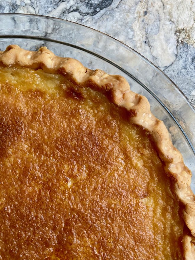 Close up of the surface of a freshly baked Tyler pie, with crimped crust in glass pie pan on marble surface. Golden brown and delicious-looking!