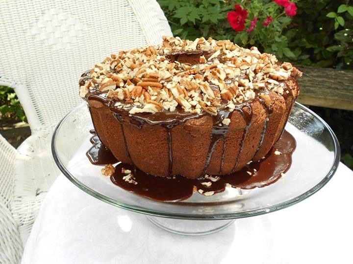 Chocolate pound cake on a glass cake stand, with chocolate glaze on top, covered with chopped pecans, on a white tablecloth outdoors, with red flowered bushes in the background