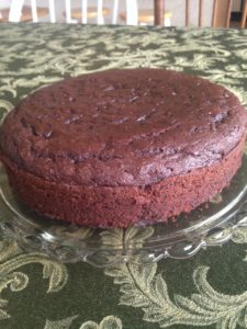 Single layer chocolate cake from the side and above, on a glass cake plate with olive-green tablecloth beneath ready to be iced.