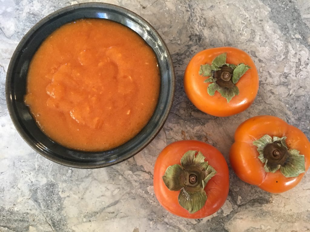 A black bowl full of bright orange persimmon puree, with three fuyu persimmons to the right all on a marble countertop