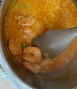 close up of red curry cooking in pot, with spoon scooping up one delicious shrimp from the sauce
