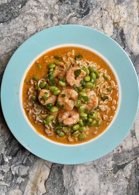 Red curry shrimp over instant ramen with edamame beans and green onion garnish in sky blue bowl centered on marble surface