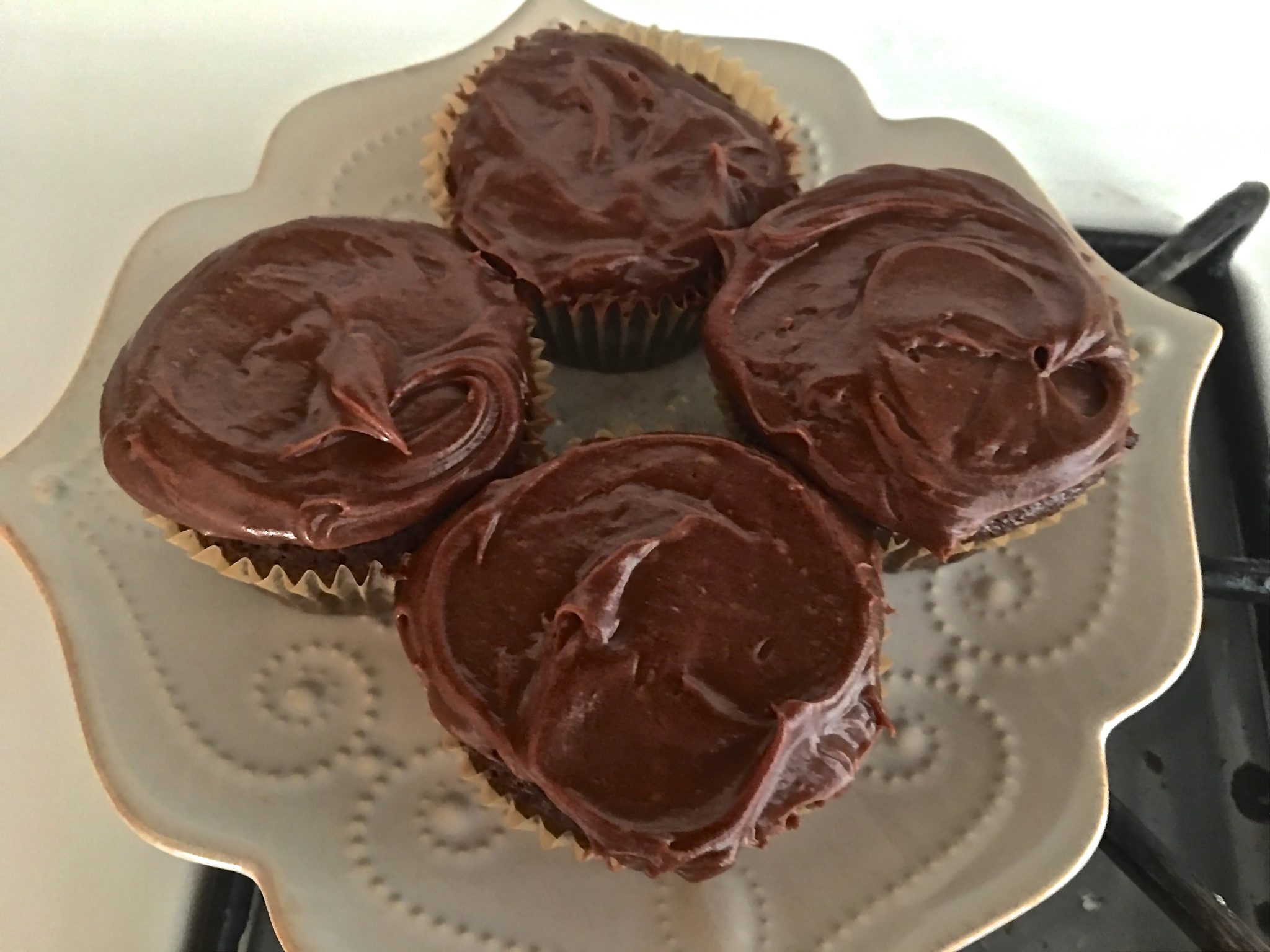 Four rich chocolate iced cupcakes in paper cupcake liners on white plate with raised curlycue design