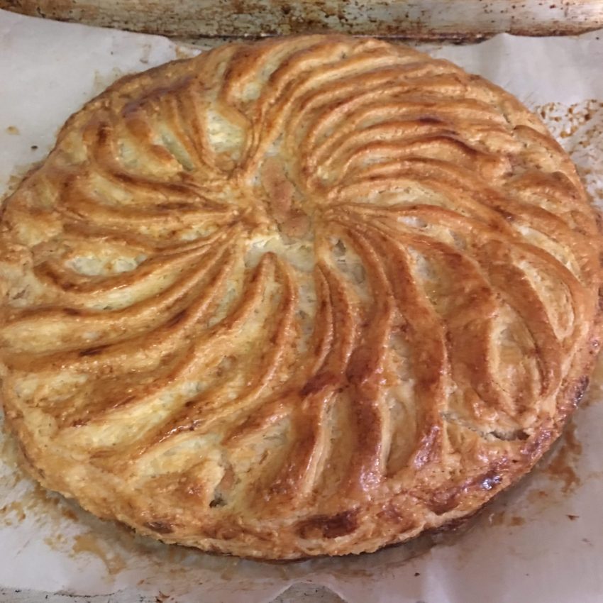 Pastry-wrapped almond custard filling with a curving designed top is galette des rois on a parchment sheet ready to cut and enjoy