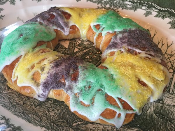 Side view from slightly above of baked and iced King Cake, glazed and decorated with three alternating wide stripes of each mardi gras colors, purple, gold, and green. 