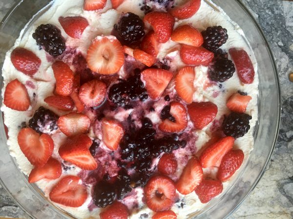 Next to last layer of trifle: Whipped cream covered with chopped strawberries and whole blackberries in syrup, over layer of smoothed whipped cream, in big glass bowl on a marble slab