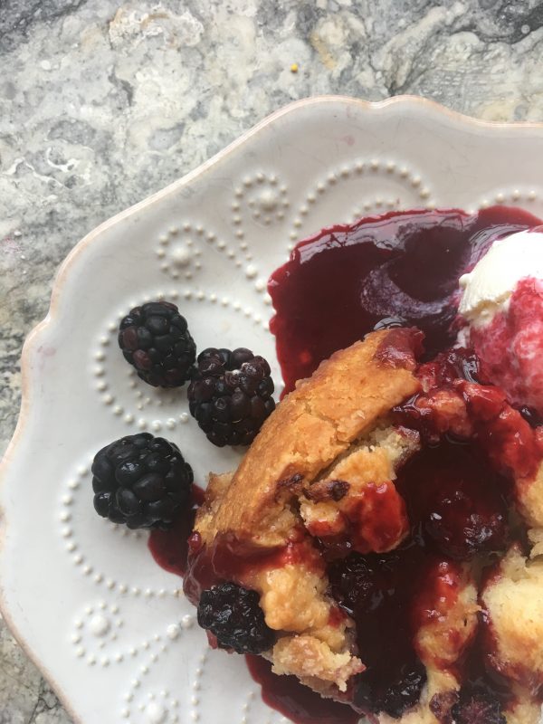 Blackberry Roly Poly, like a cobbler with biscuity dough, brown sugar and butter, on a white plate with raised dots, blackberries, a little ice cream and juice on a marble slab