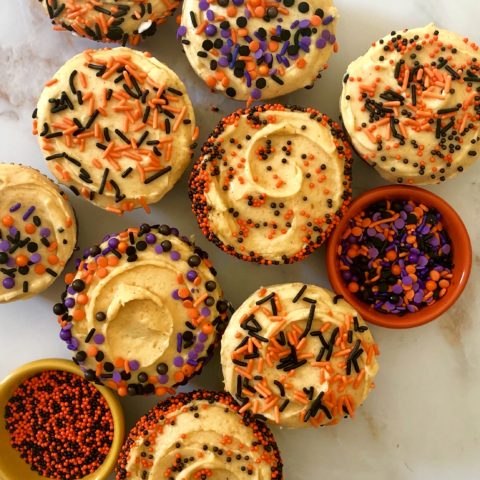 Close up of pumpkin spice cupcakes with pumpkin frostingm, top down, with bowls of spooky sprinkles, on a marble surface