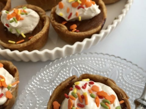 Looking down on an array of Pumpkin Pie Minis topped with whipped cream and sprinkles; glass pate with bubbly design and ceramic tart pan decorate the image.