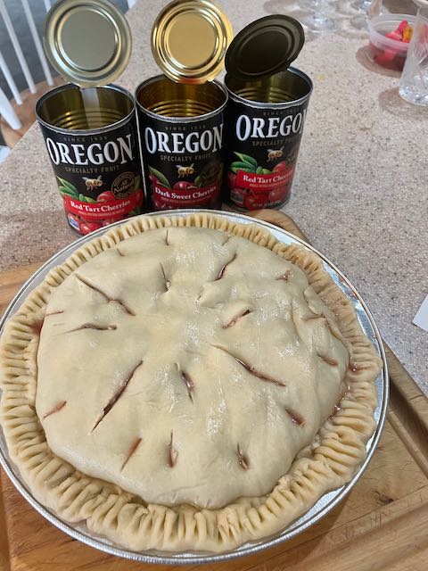 three cans of sour and dark cherries, open, next to unbaked cherry pie with double crust, steam vents, and fork-tine crimping on crust edge