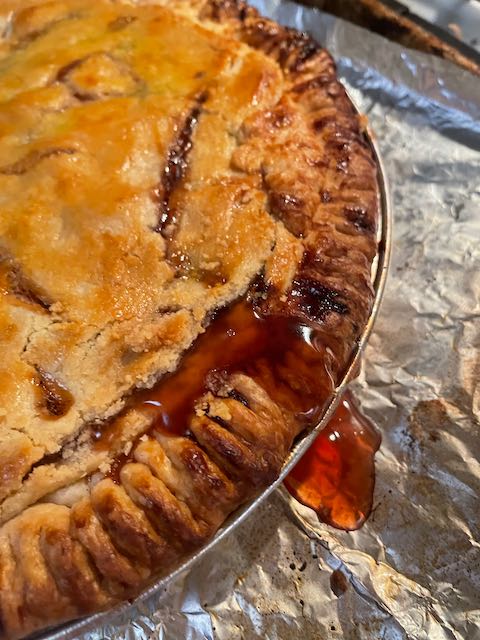 close up of half the baked pie on a foil pan to catch syrup that bubbles out as it bakes; bright red syrup and golden crust with fork-tined crust