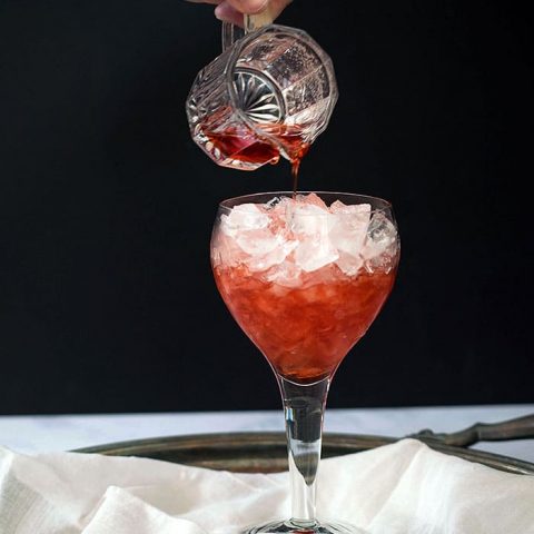 Stemmed wine glass of crushed ice with small pitcher pouring red liquid over ice ; white silk under glass, black background
