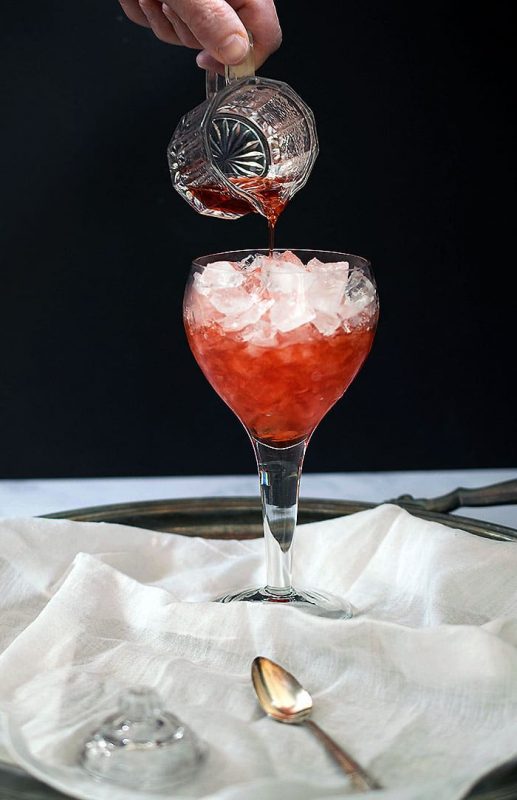 Red syrup (strawberry shrub) being poured from small glass pitcher overhead into wine glass full of crushed ice, placed on white silk fabric against black background
