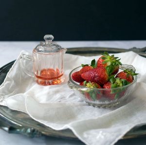 Shrub in petite glass pitcher with top, next to small glass bowl of whole berries, on white linene , on a tray on a marble table with black background in the distance