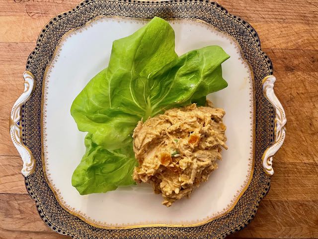 Individual serving of chicken salad on one lettuce leaf centered on elegant gold trimmed square plate set on wooden board.