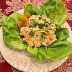 Curried chicken salad served on Boston lettuce leaves atop white plate on red brocade tablecloth apricots and herb garnishes